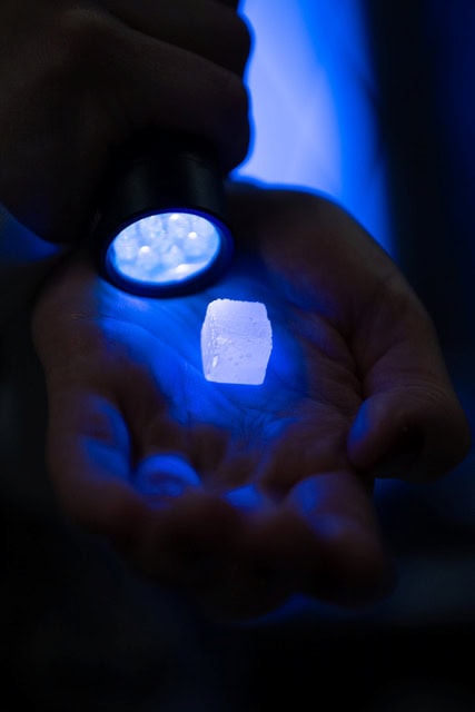 A close up of a hand holding an approximately one-inch cube glowing blue from a UV flashlight held near the surface.