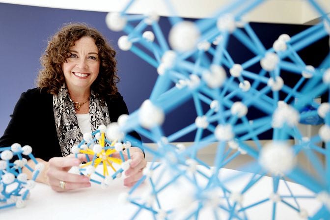 Sharon Glotzer sits at a round, white table. She holds a ball-and-stick model of a crystal. Two similar models are on the table in front of her.
