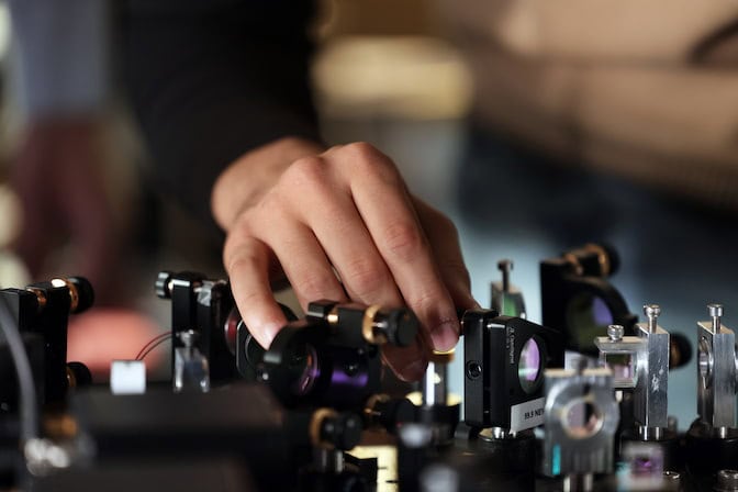 The optical table is crowded with small lenses and mirrors. A hand reaches among them.