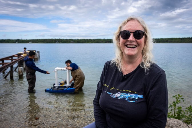 Beth Crosswhite smiles and sits at a stone beach. The bright blue waters of Lake Michigan behind her. In the water behind her, two men examine a catamaran-shaped device.