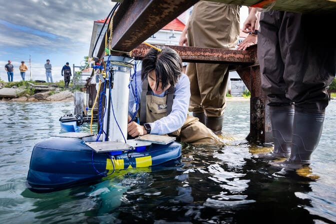 A man crouches below the steel railing of a dock. Waist deep in lake water, he handles wires on a buoy, which resembles a blue inner tube around a metal post. The inner tube portion of the device sports the Michigan block M.