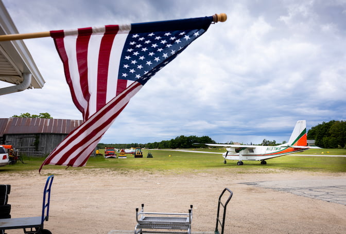 A runway cuts through a grass lawn behind the flag. A white plane is parked near the runway. The plane has an orange stripe that stretches the entire length of the body and gets thicker toward the tail. Green stripes flank each side of the orange stripe.