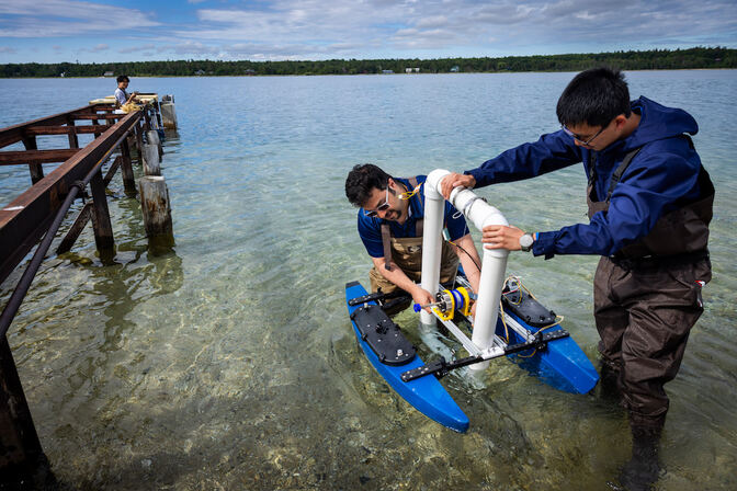 Two researchers are holding a device that looks like a blue catamaran built around a square, PVC pipe frame. A small LED connected at the top of the frame is on. Another man stands at a dock further away. All the researchers are wearing waders and standing in the lake. Two researchers are holding a device that looks like a blue catamaran built around a square, PVC pipe frame. A small LED connected at the top of the frame is on. Another man stands at a dock further away. All the researchers are wearing waders and standing in the lake.