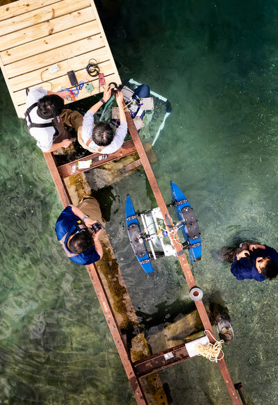 Four researchers stand at a dock with large gaps in the boardwalk providing access to the water underneath. Two devices are tied to the dock. A researcher connects a wire extending from one of the devices to a circuit board resting on the boardwalk.