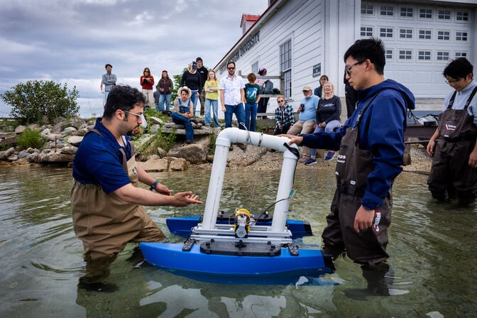 Two men stand knee deep in a lake. One of the men holds a device that looks like a blue catamaran built around a square, PVC pipe frame. The other man gestures toward the device. A crowd of people watch from the shore nearby.