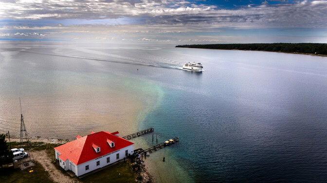 An aerial view of a white ferry approaching Beaver Island. The boathouse has a vibrant, scarlet-colored roof.