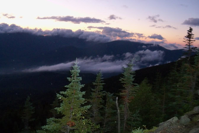 A sunrise landscape in the mountains. Trees are visible in the foreground and blue mountains in the background with long, streaking clouds in the sky.