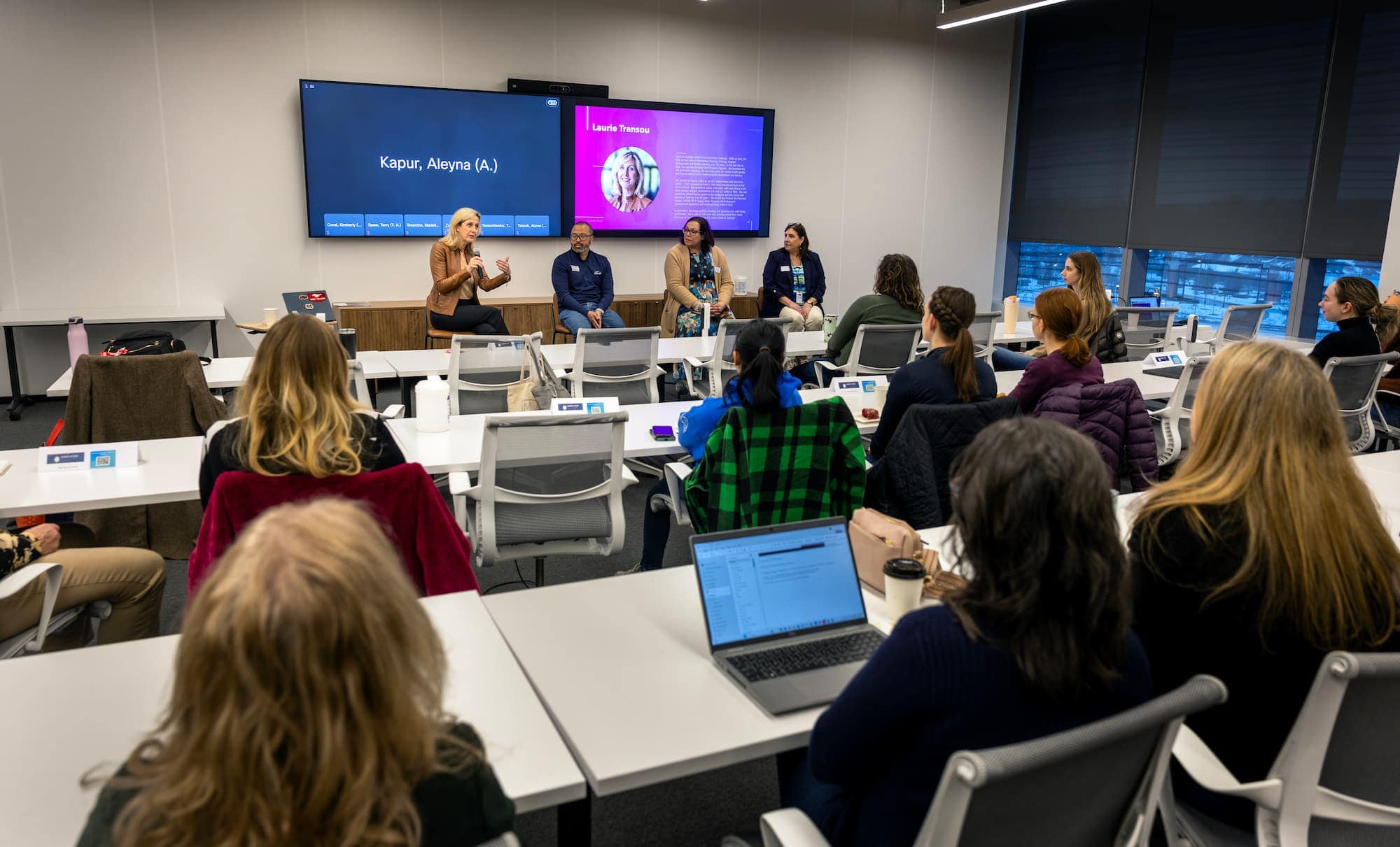 A seminar panel featuring four speakers in a modern classroom, with an audience attentively listening. One speaker gestures as she addresses the crowd, and presentation screens display information about the speakers.