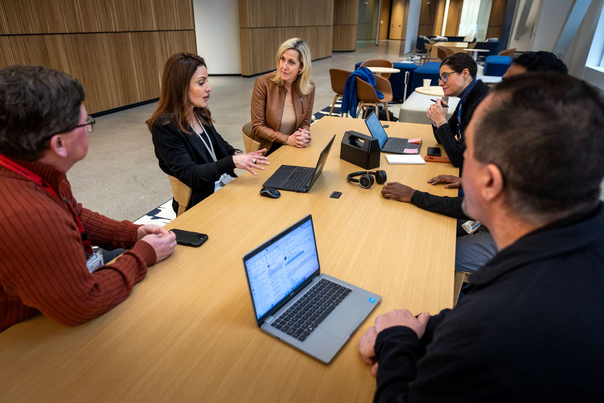 A meeting at the Ford World Headquarters with five team members gathered around a wooden table, discussing. Laurie Transou, in a brown leather jacket, is among them.