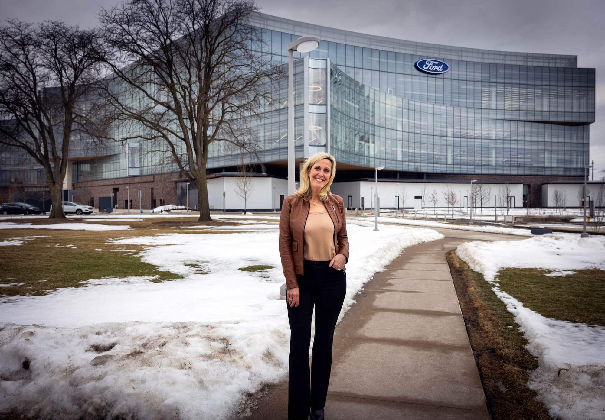 Laurie Transou stands smiling in front of the modern Ford World Headquarters, wearing a brown leather jacket and black pants, with patches of snow and bare trees around her.