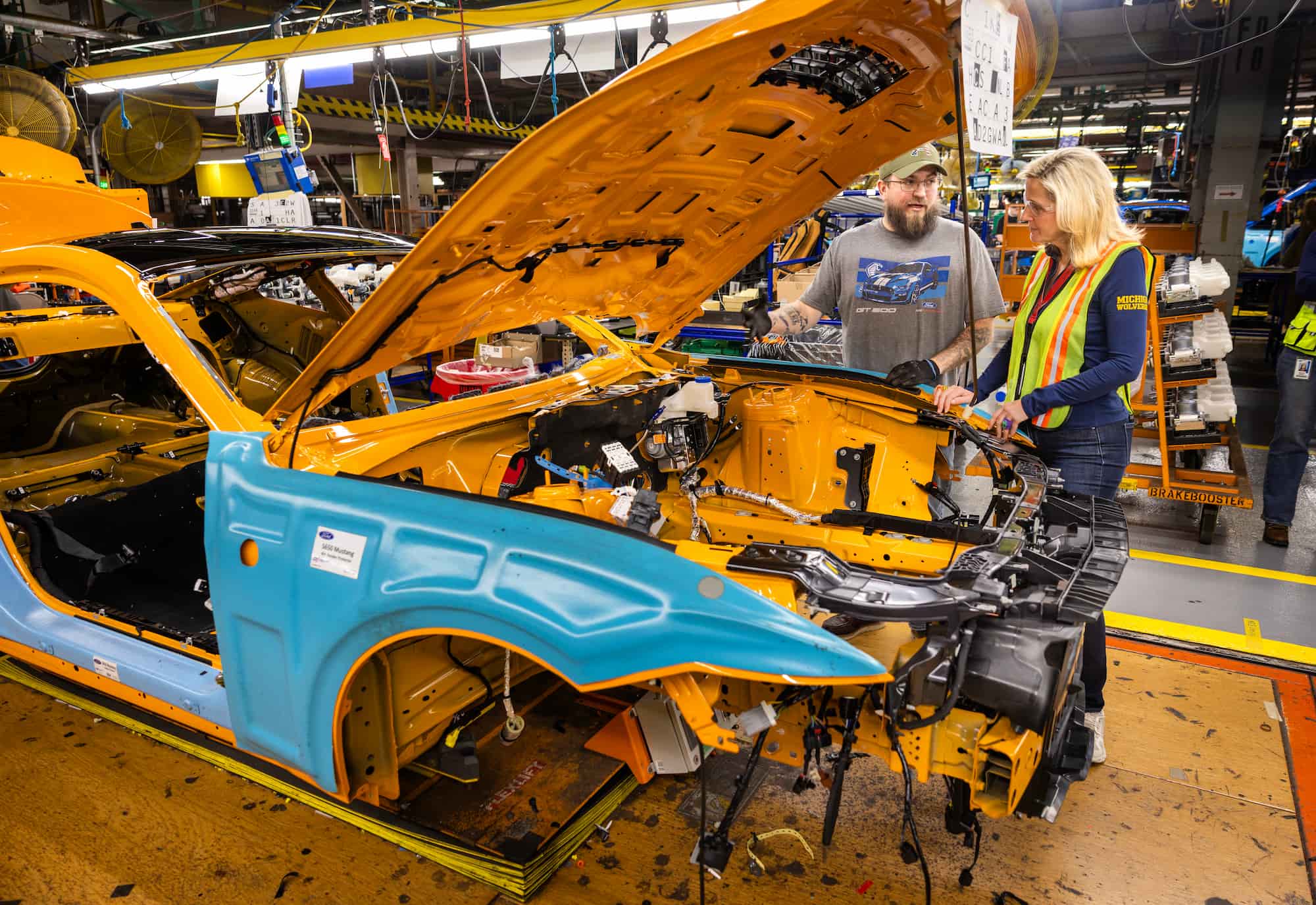 A partially assembled Mustang body moves through the production process at the Flat Rock Assembly Plant, surrounded by machinery and workers.