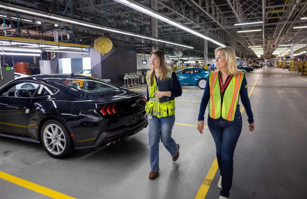 Ford employees wearing safety vests walk by a vehicle on the Mustang car at the Flat Rock Assembly Plant.