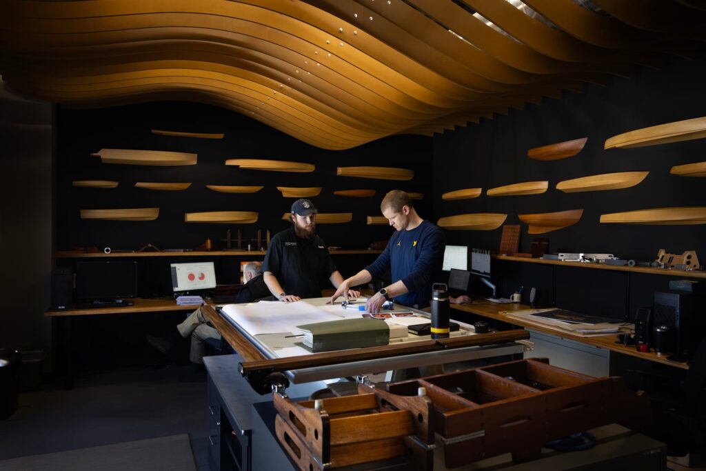Two men discuss design plans inside a workshop-like space filled with wooden boat hull models mounted on the walls. 