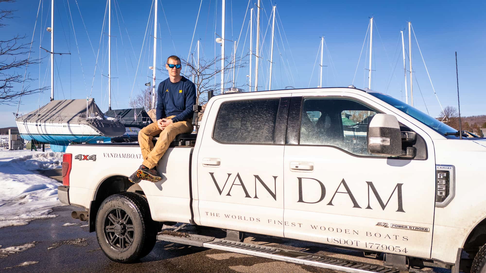 Ben Van Dam sits on the bed of a white Van Dam Boats pickup truck at the Boyne City Marina, with sailboats and snow-covered docks in the background. 