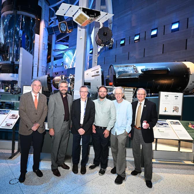 Five men stand beneath a model of a spacecraft, about the size of a toaster oven, which hangs from the ceiling.
