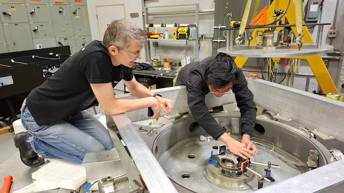 In U-M’s Plasma, Pulsed Power, and Microwave Laboratory, one researcher reaches with both hands to adjust the X-pinch load hardware in U-M’s MAIZE pulsed power facility, while another researcher looks on.