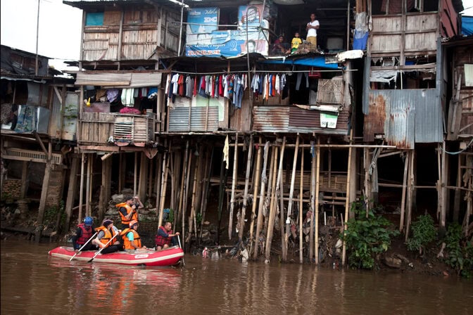 Five people, in a red inflatable raft, paddle by a house on stilts.
