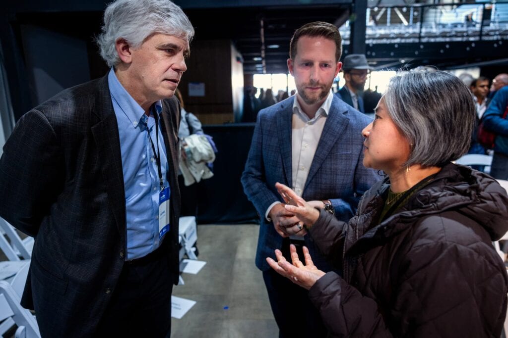 A man and a woman engage in a serious conversation while another man looks at them at an event.