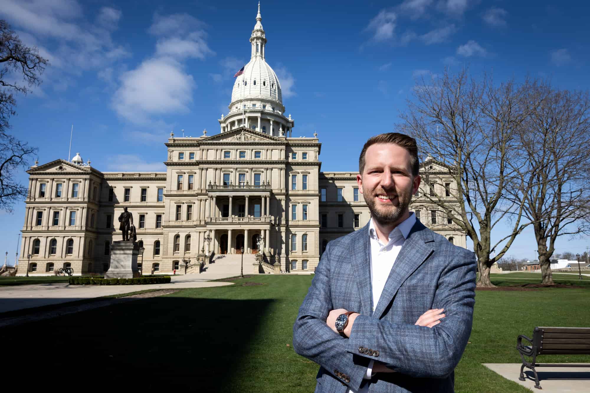 A man in a blue blazer stands with arms crossed in front of the Michigan State Capitol on a sunny day with blue skies.