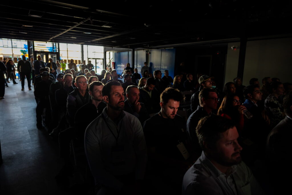 A crowd of attendees sits closely together in a darkened event space, listening to a presentation.