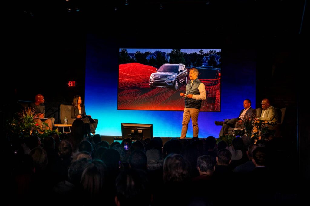 A speaker presents on stage with a large screen displaying a car image to an audience