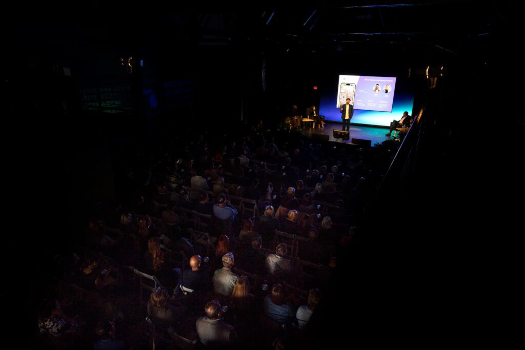 : A dimly lit auditorium filled with people watching a presentation in front of a large stage screen.