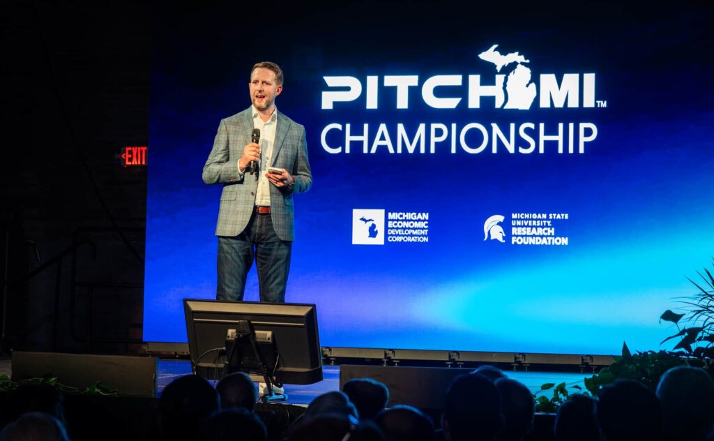 A man speaks on stage in front of a large screen reading “PitchMI Championship.”