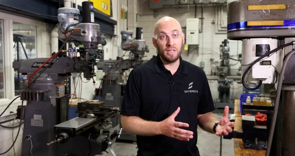 A man in glasses speaks and gestures while standing in an industrial shop. 