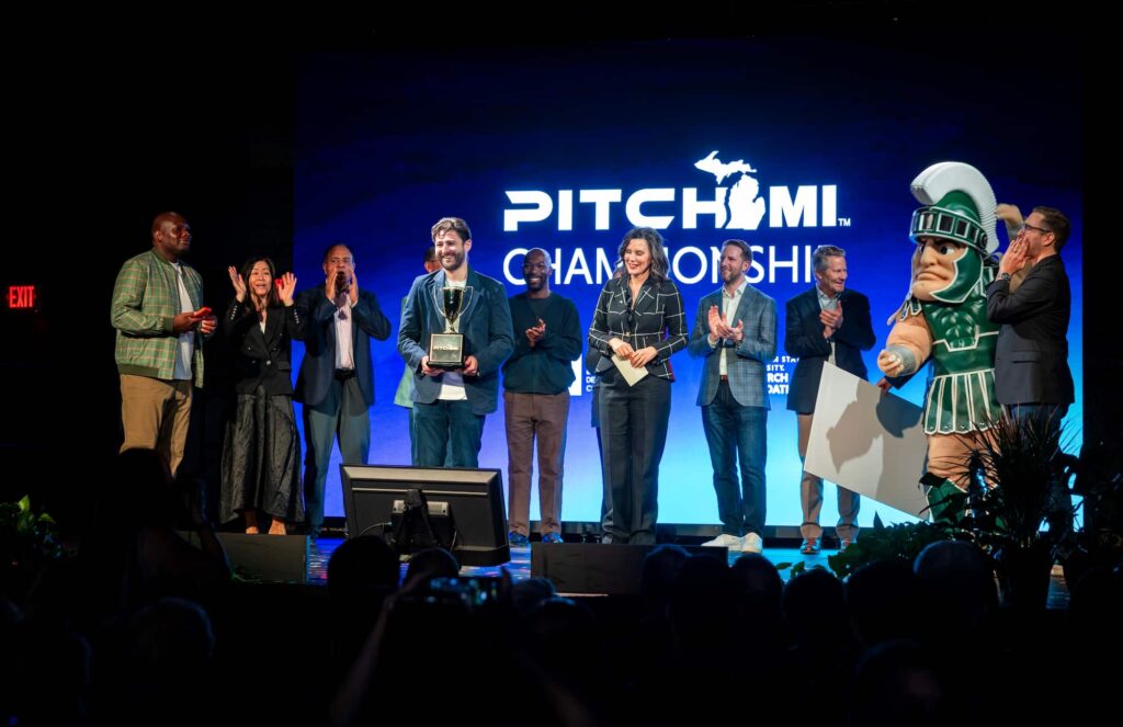 A group of people stand on stage during the PitchMI Championship event, with a MSU mascot visible at the side.