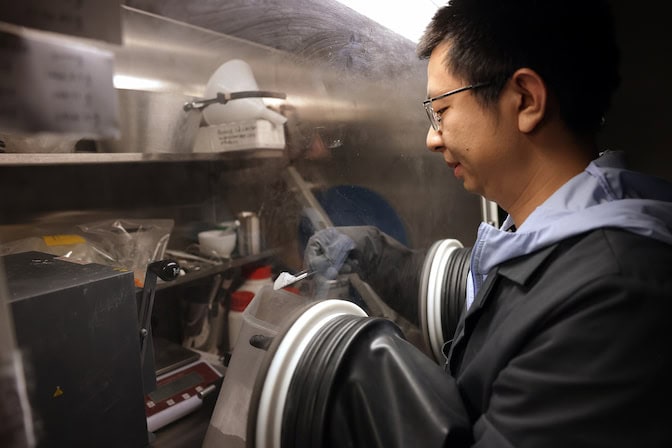 A man stands outside of a glass laboratory enclosure with his hands pushed through two circular openings with black gloves. Inside the ox, he holds a plastic container with one hand and a metal scooper holding salt in the other.
