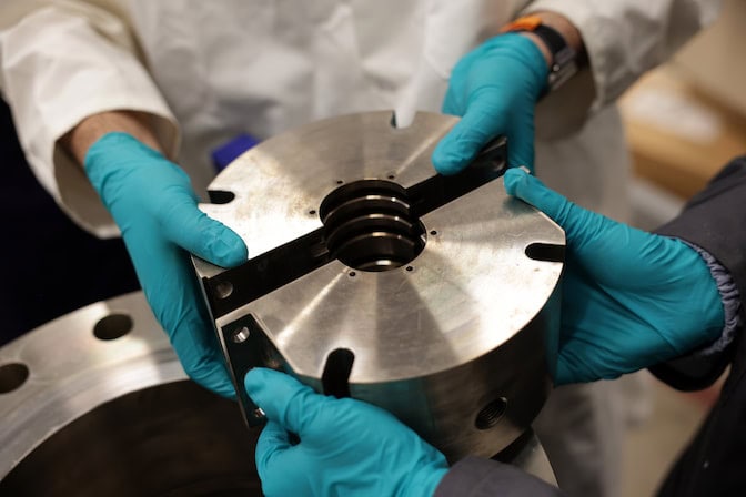 A closeup of two pairs of gloved hands each holding a half of a cylindrical shaft seal with an opening in the middle for the shaft to go through.