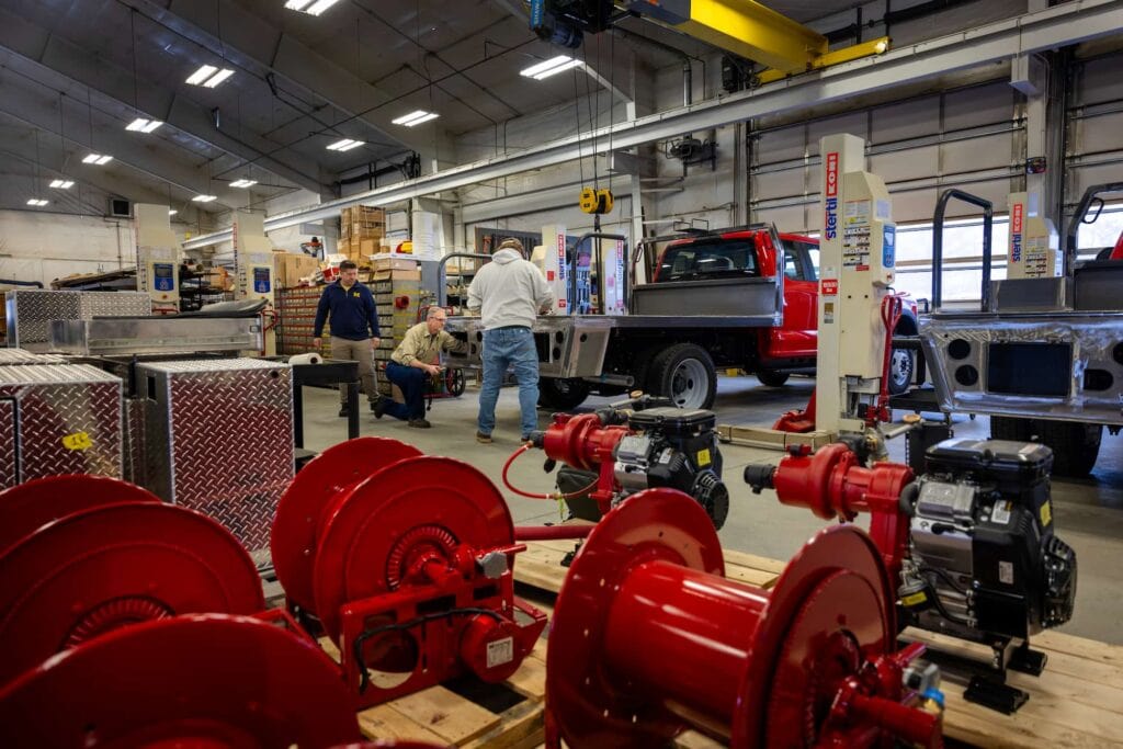 A wide view of a workshop with fire truck components, tools, and equipment laid out on the floor with the people behind it.