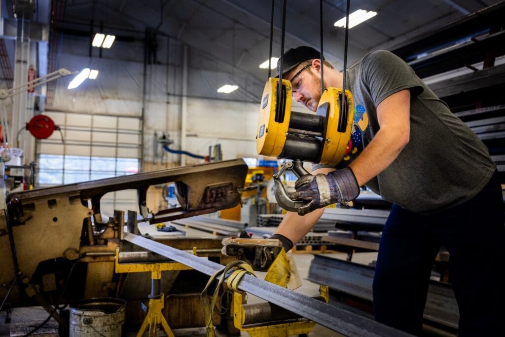 A worker uses an overhead hoist to position a steel beam onto a metal-cutting machine inside a fabrication shop.