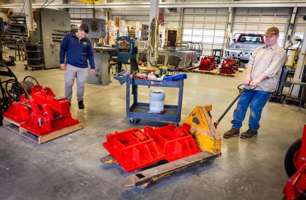 Two men move heavy red steel machine components with a pallet jack across the floor of a manufacturing shop at the Forest Fire Experiment Station.