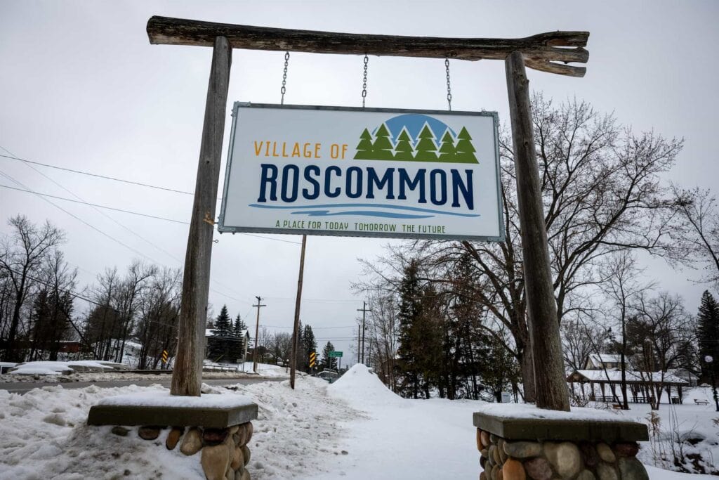 A wooden roadside sign reading “Village of Roscommon” welcomes visitors at the entrance to the town.