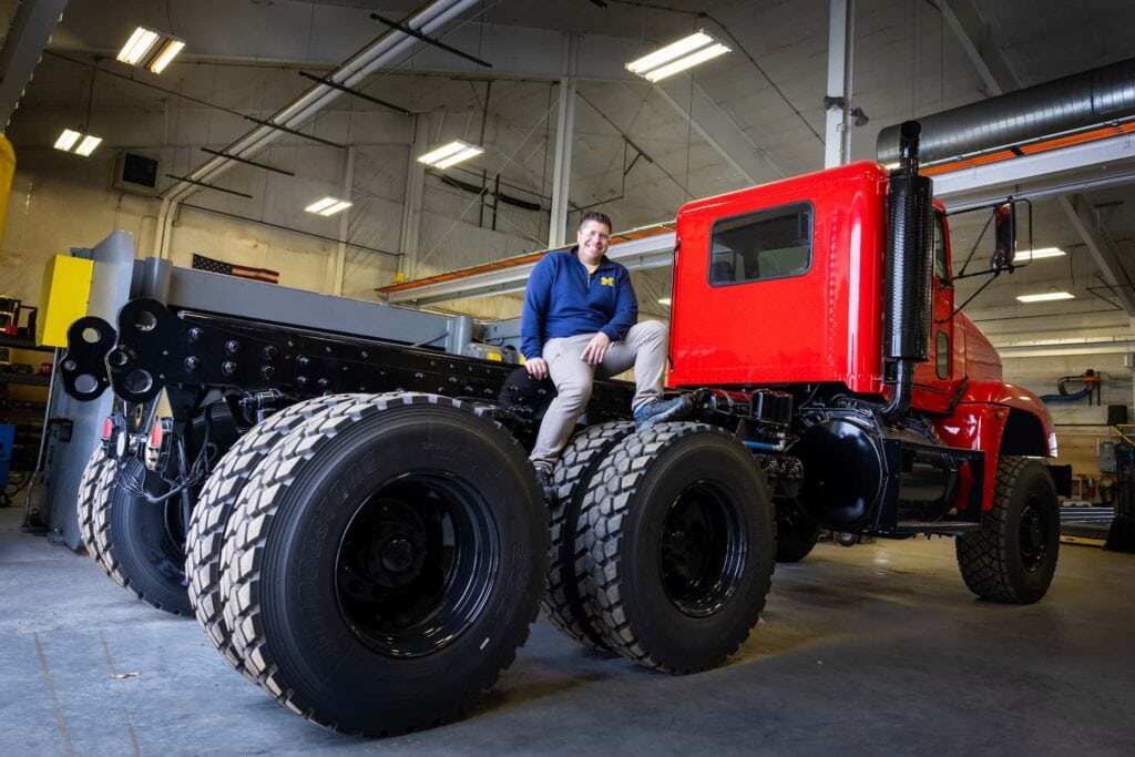 A man sits on the bed of a partially assembled red fire truck inside a workshop.