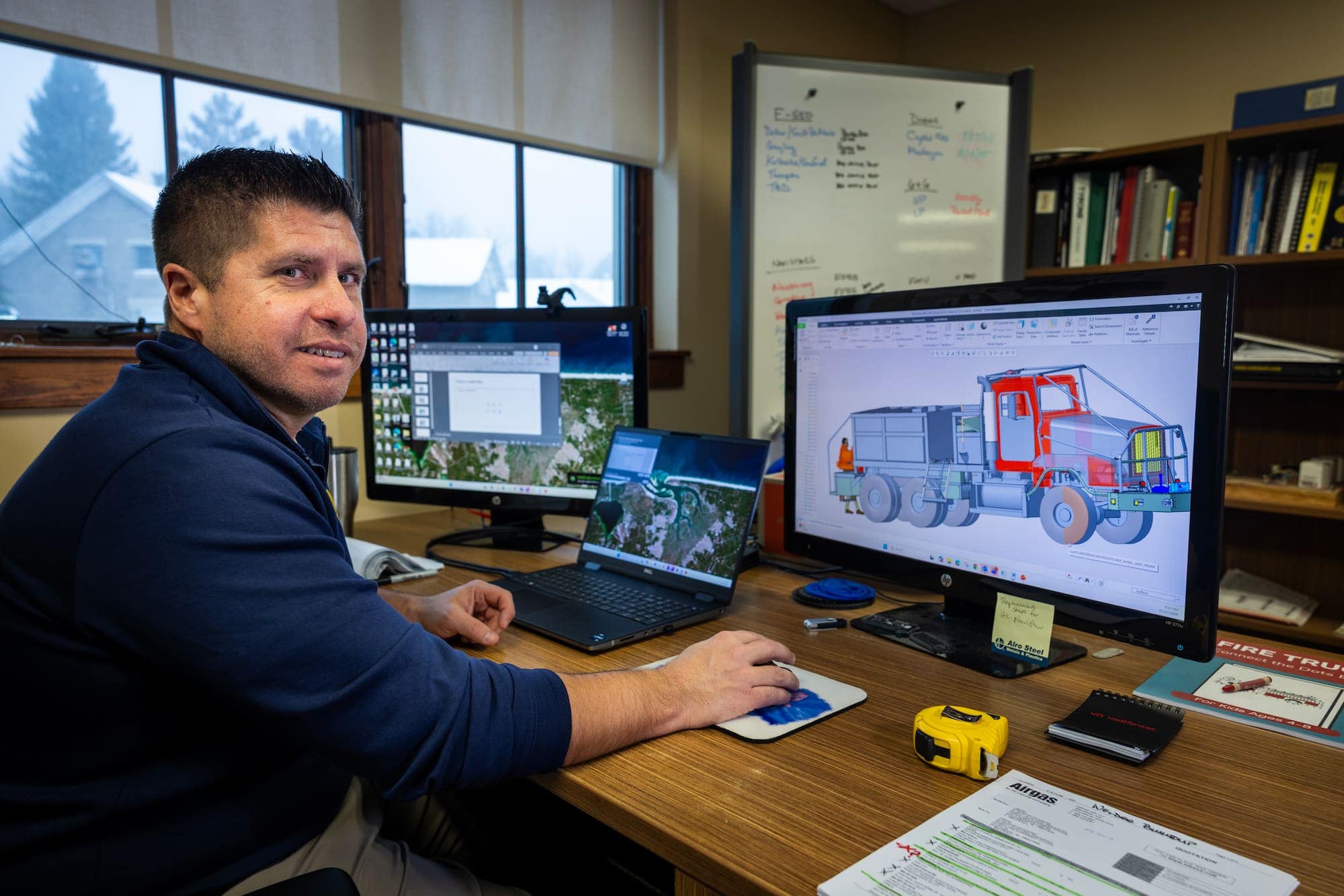 A man seated at a desk turns toward the camera while working on a computer with a fire truck design on screen.