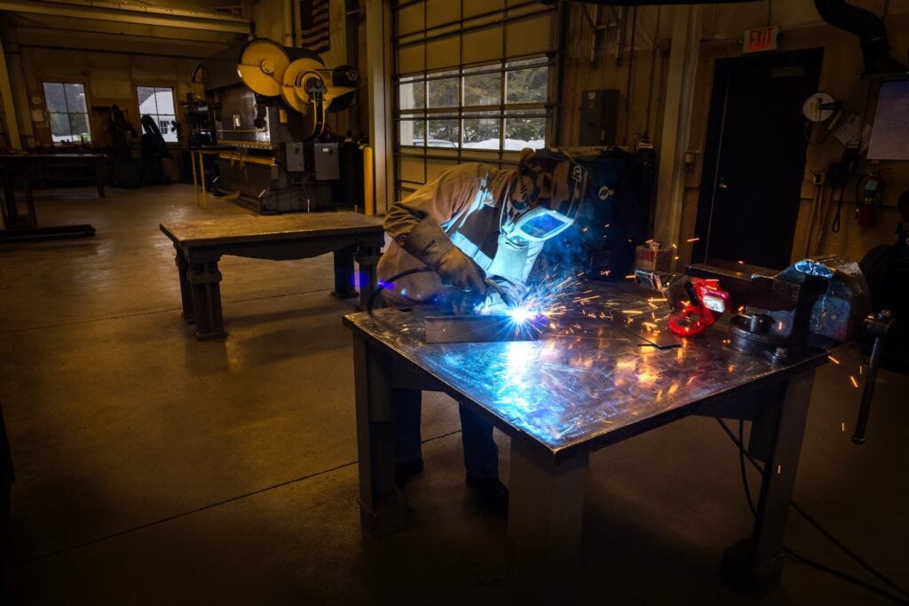 Welder works at a metal table inside a workshop with sparks flying.