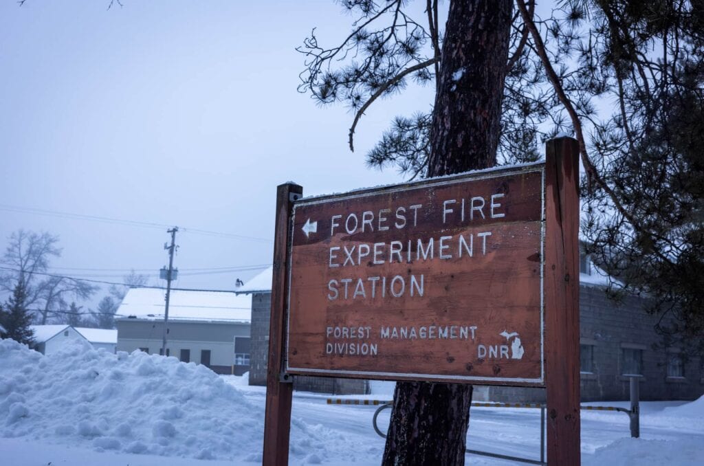 A wooden sign reading “Forest Fire Experiment Station” stands outdoors in winter, surrounded by snow and trees outside of the DNR’s Forest Fire Experiment Station.