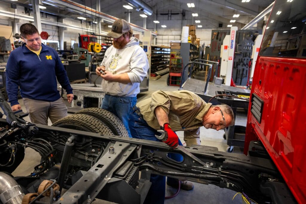 Three people work together on the exposed chassis of a fire truck inside a large workshop.