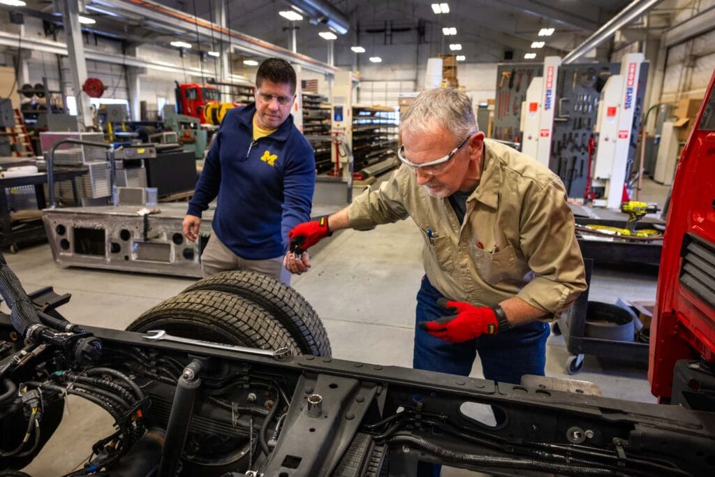 Two technicians inspect and work on a fire truck chassis using tools inside an industrial space.
