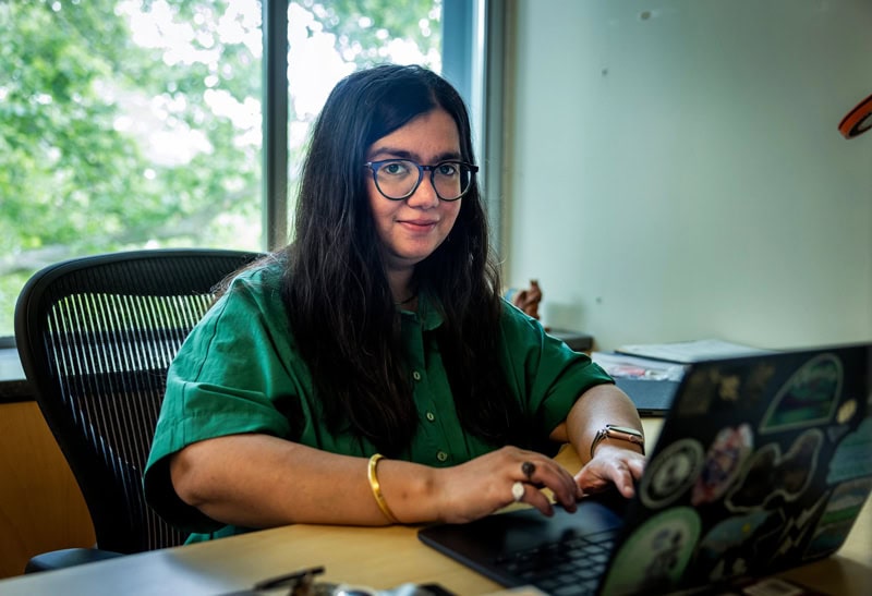 A woman sits in an office in front of a laptop computer, looking directly at the camera.