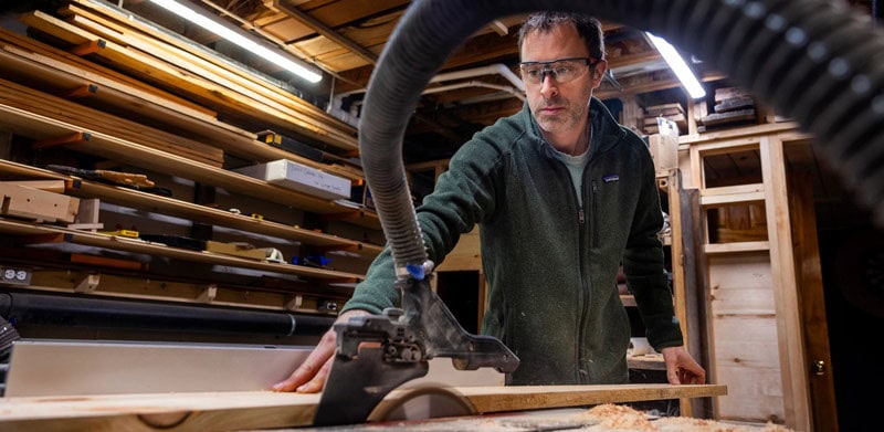 Pete Cook operates a woodworking machine in a workshop, guiding a piece of wood while wearing protective glasses.