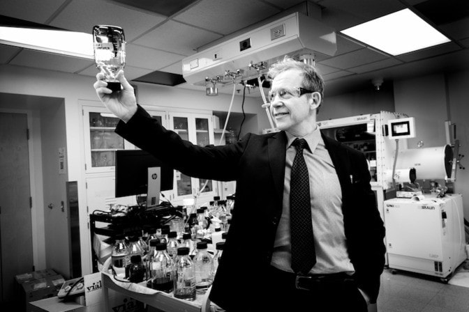 A man in a suit holds up a bottle with some chemicals in a lab setting. 