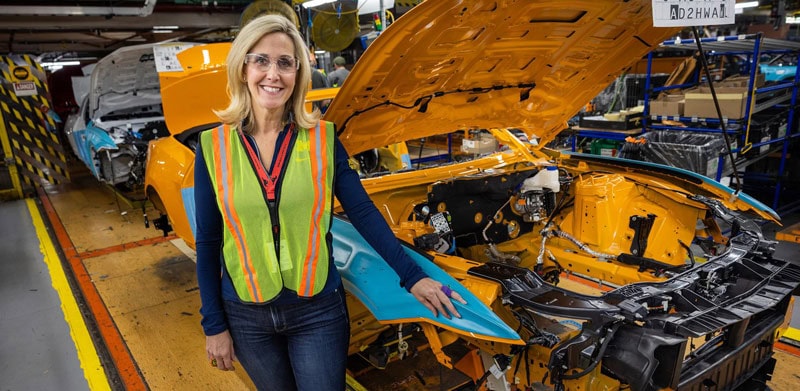 Laurie Transou wears a safety vest and stands beside a partially assembled vehicle in a manufacturing facility.