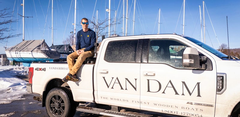 Ben Van Dam sits on a white pickup truck at a marina, with sailboats and a clear blue sky in the background.