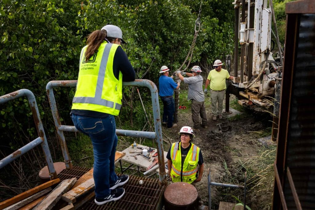 Researchers and crew members gather around drilling equipment at an outdoor field research site.