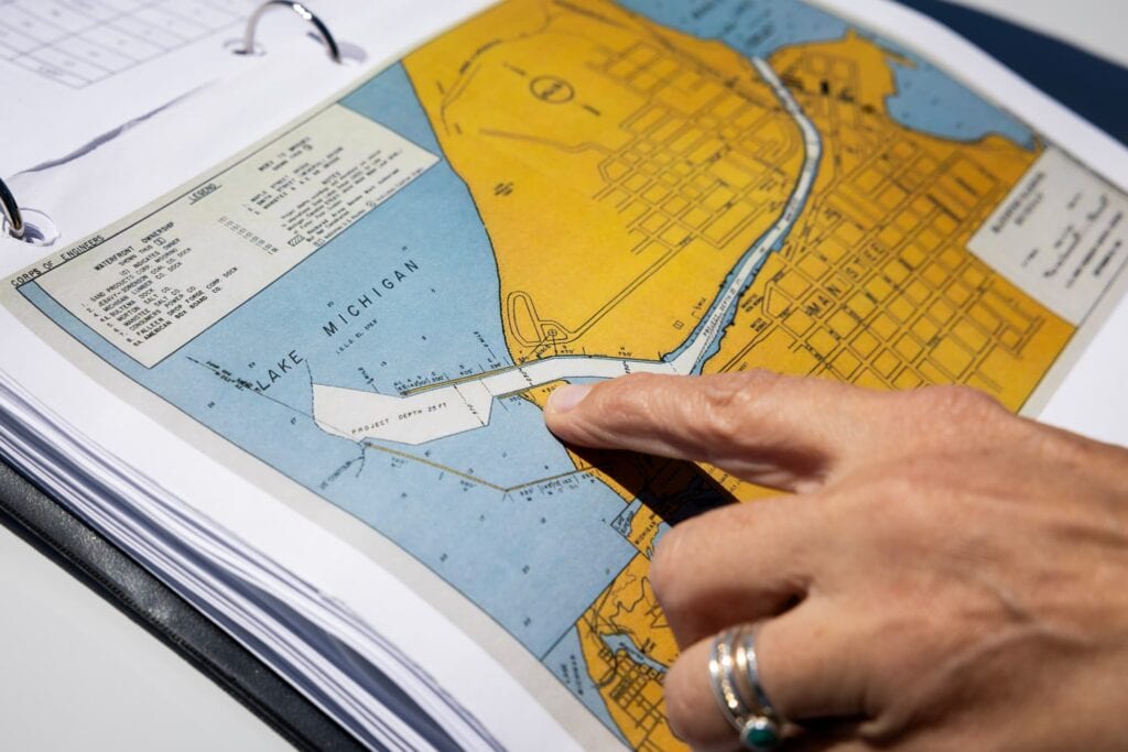 A hand pointing to a printed coastal map and engineering plans on a desk.