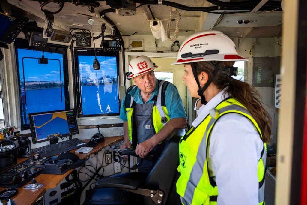 Two workers wearing hard hats talk inside a boat's wheelhouse.