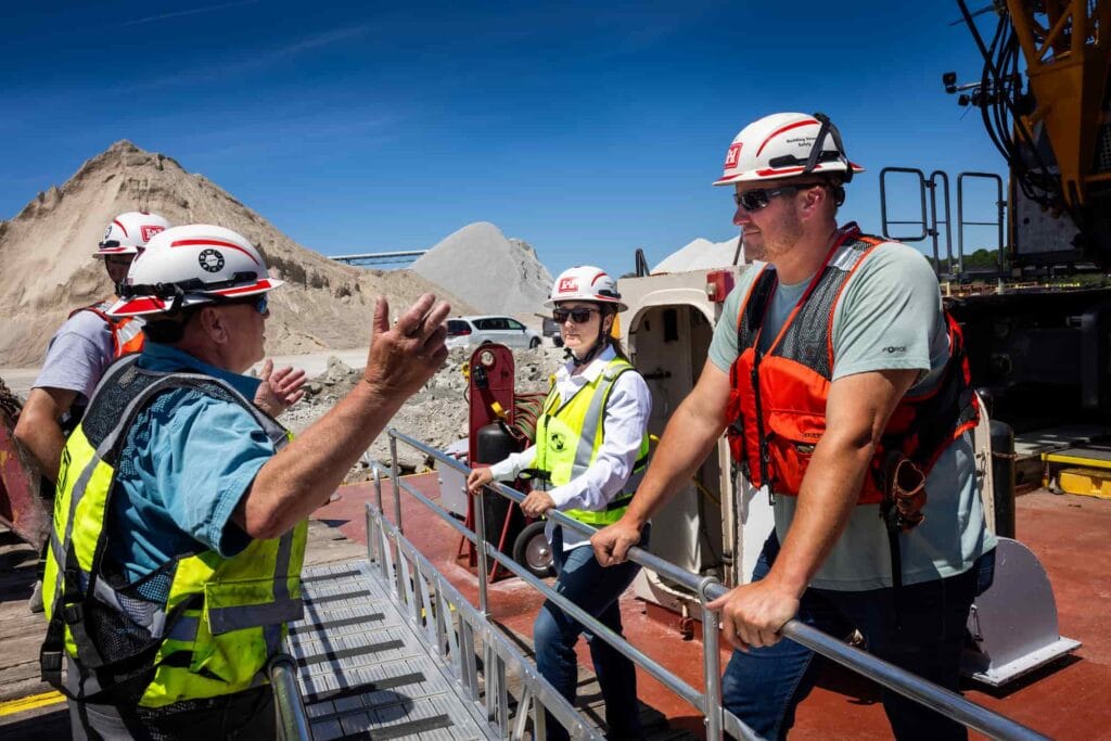 Workers in hard hats and safety vests talk aboard a workboat at a dock.
